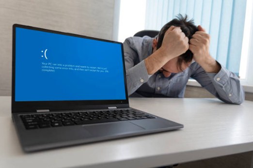 Frustrated man with his head in his hands next to a laptop showing a blue screen error